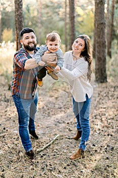 Happy young parents and kid little son, walking together outdoor in autumn forest or park, holding their baby on hands