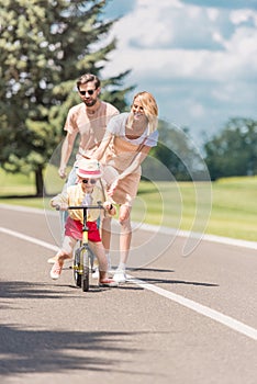 happy young parents looking at little daughter riding bicycle