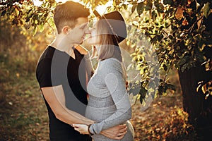 Happy young parents, mom and dad, hugging baby bump, enjoying beautiful moment at sunset. Stylish pregnant couple holding hands on