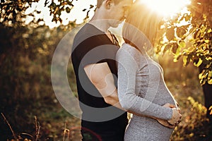 Happy young parents, mom and dad, hugging baby bump, enjoying beautiful moment at sunset. Stylish pregnant couple holding hands on