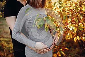 Happy young parents, mom and dad, hugging baby bump, enjoying beautiful moment at sunset. Stylish pregnant couple holding hands on