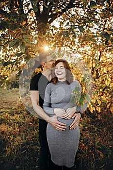 Happy young parents, mom and dad, hugging baby bump, enjoying beautiful moment at sunset. Stylish pregnant couple holding hands on