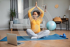 Happy young pregnant black woman with big belly sits in lotus position practices yoga and watches video workout