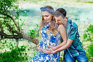 Happy and young pregnant couple hugging in nature on sunset. New family expecting baby in scenic place near river in summertime.