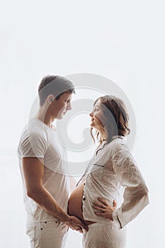 Happy young pregnant couple in white pajamas holding belly bump in morning light. Stylish pregnant family, mom and dad, relaxing