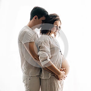 Happy young pregnant couple in white pajamas hugging and holding belly bump in morning light. Stylish pregnant family, mom and dad