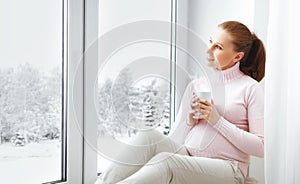 Happy young pregnant woman with cup of tea on window