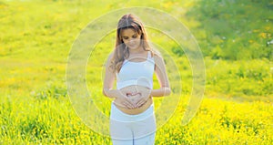 Happy young pregnant woman makes heart sign on her belly with her hands in summer park