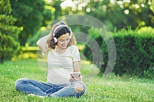 Happy young pregnant woman sitting on carpet and listening music on headphones from smartphone at park. Pregancy, Relaxation and