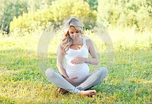 Happy young pregnant woman sitting on grass doing yoga in summer