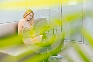 Happy young pregnant woman is sitting in hallway of hospital waiting to see gynecologist.