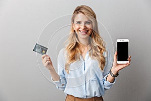 Happy young pretty blonde business woman posing  grey wall background holding credit card showing display of mobile phone