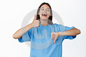 Happy young pretty girl laughing, showing thumbs up and thumbs down, like or dislike, standing in blue t-shirt over