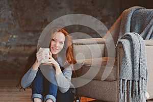 Happy young readhead woman drinking hot coffee or tea at home. Calm and cozy weekend in winter