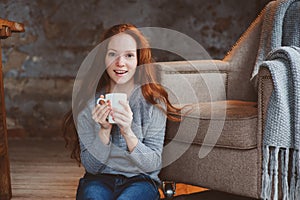 Happy young readhead woman drinking hot coffee or tea at home. Calm and cozy weekend in winter