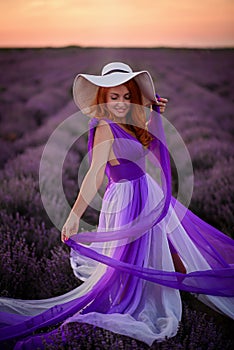 Happy red-haired woman in luxurious dress standing in lavender field at sunset