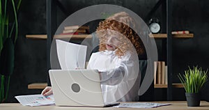 Happy young redhead business woman professional talking on phone while working with laptop computer sitting at office