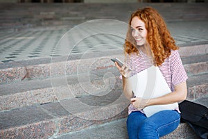 Happy young redhead girl holding phone and smiling, sitting on the stairs outdoors