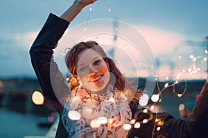 Happy young redhead woman playing with fairy lights outdoors and