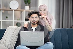 happy young relaxed Muslim couple working on laptop pc at cozy home interior. Handsome man sitting on sofa with laptop