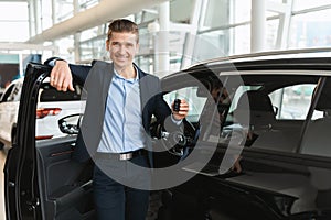 Happy young salesman standing near open car, showing auto key, selling new vehicles at modern dealership, empty space