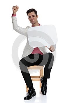 Happy young seated businessman holding a speech bubble celebrates success