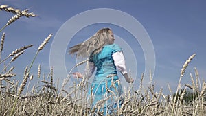 Happy young slender woman with a long fair hair in a blue dress rotates in the field of ripe wheat in summer sunny day