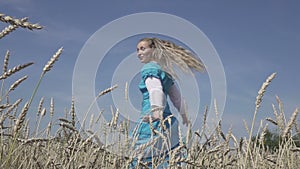 Happy young slender woman with a long fair hair in a blue dress rotates in the field of ripe wheat in summer sunny day.Slow motion