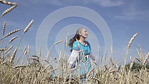 Happy young slender woman with a long fair hair in a blue dress rotates in the field of ripe wheat in summer sunny day.Slow motion