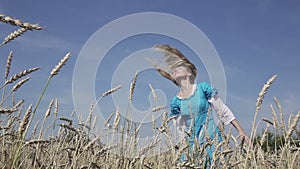 Happy young slender woman with a long fair hair in a blue dress rotates in the field of ripe wheat in summer sunny day.Slow motion