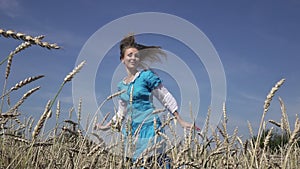 Happy young slender woman with a long fair hair in a blue dress rotates in the field of ripe wheat in summer sunny day.Slow motion