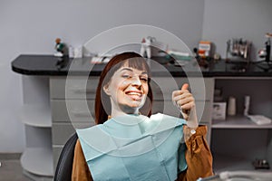 Happy young smiling woman showing thumb up sitting at the dentist office after dental examination and painless treatment