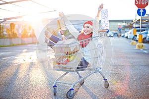 Happy young smiling woman sitting in shopping cart with a lot of gift boxes and two shopping bags in her hands