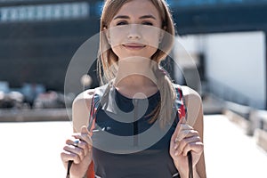 Happy young sport woman in stylish training outfit with jumping rope on her shoulders is smiling, standing outside