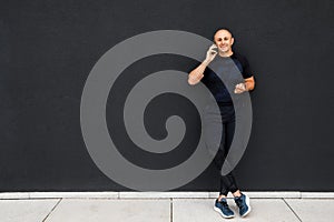 Happy young sporty man talking on phone, having break after training, standing at black wall, copy space