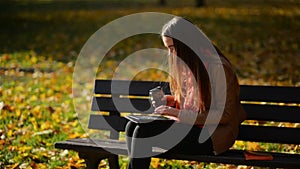 Happy Young Student Girl with a Tablet and a Disposable Coffee Cup Sitting on the Bench and Using Laptop in a Autumn