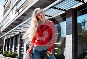 Happy young stylish girl in leather jacket and jeans looking in camera walking around city street.beautiful blonde with long hair