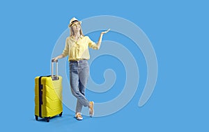 Happy young tourist with suitcase presenting something on blue copy space background