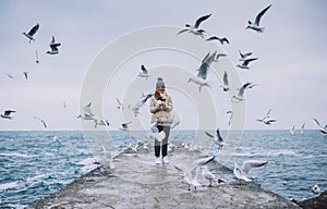 Happy young tourist woman feeds seagulls on the sea. Pretty female wearing coat, scarf and watching flying seagulls by the sea in