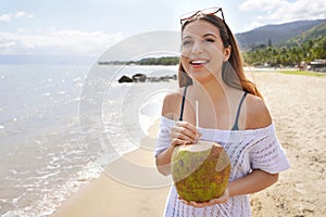 Happy young tourist woman holding green coconut on tropical beach. Summer vacation concept