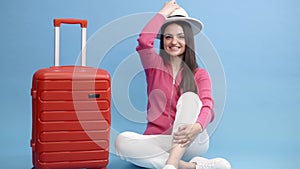 Happy young tourist woman sitting at the suitcase and going to travel on holidays isolated on blue background