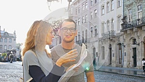 Happy young tourists couple holding a paper map of ancient European city early in the morning on empty square and