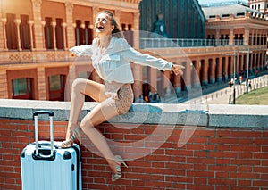 Happy young traveller woman rejoicing while sitting on parapet