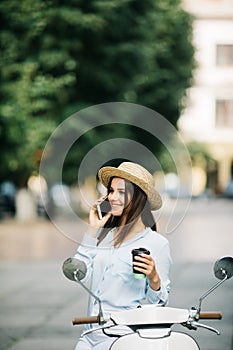 Happy young woman drinking takeaway coffee near her moped and talking phone on city street