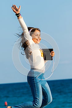 Happy young tween girl jumping with headphones and tablet on beach