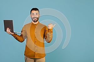 Happy young unshaved man standing with laptop and pointing aside at free space over blue background