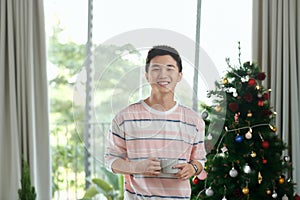 Happy young Vietnamese businessman standing near fir tree, holding coffee cup