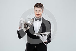 Happy young waiter lifting metal cloche from serving tray