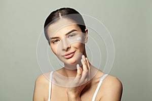 Happy young woman applying moisturizing cream on her face. Photo of pretty spa model with clean skin on light green background.