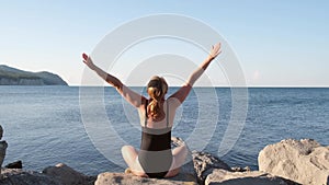 Happy young woman arms outstretched on the beach at sunrise. Girl meditating in a black swimsuit in the early morning